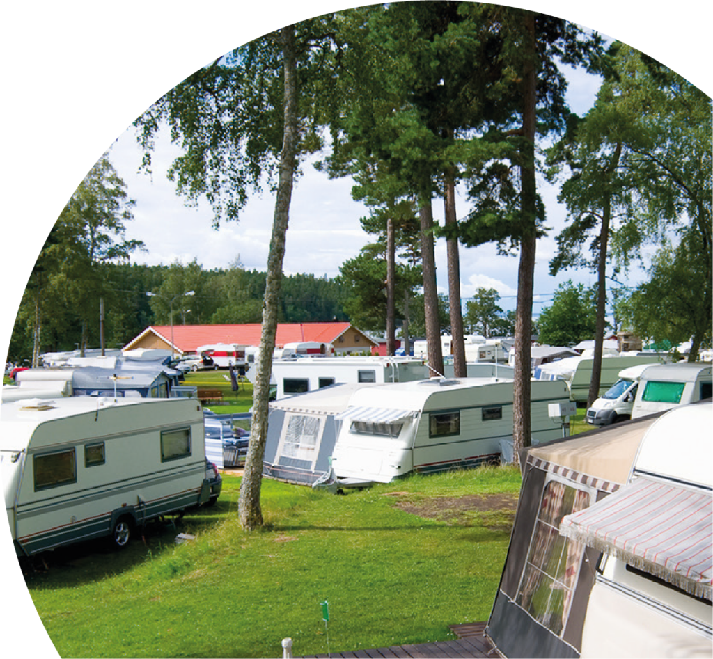 Campingplats fylld av husvagnar, träd, gräsmatta och ett hus i bakgrunden.