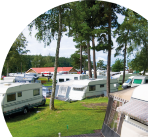 Campingplats fylld av husvagnar, träd, gräsmatta och ett hus i bakgrunden.