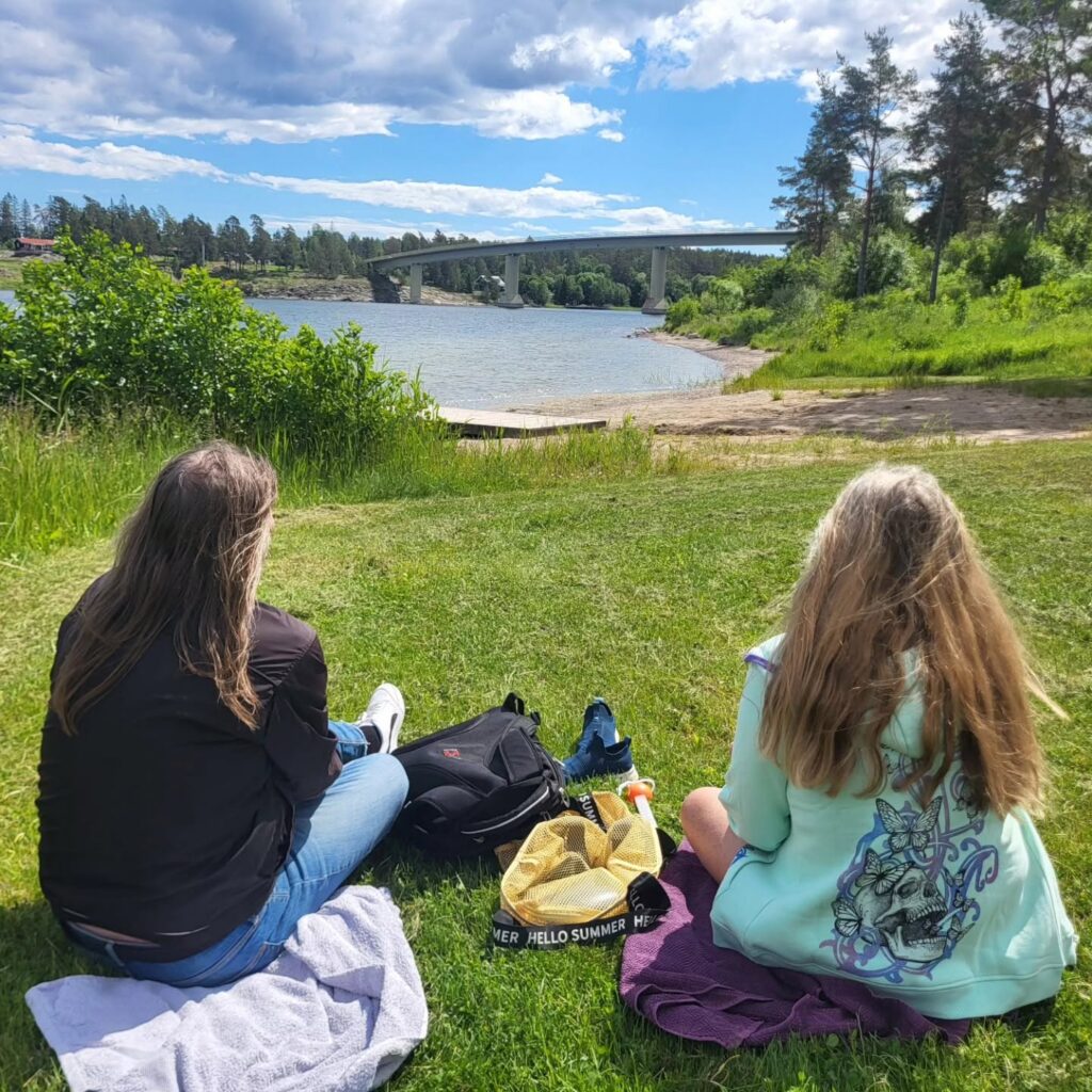 Två kvinnor har picknick ovanför Bergby badplats med Väddöviken i bakgrunden.